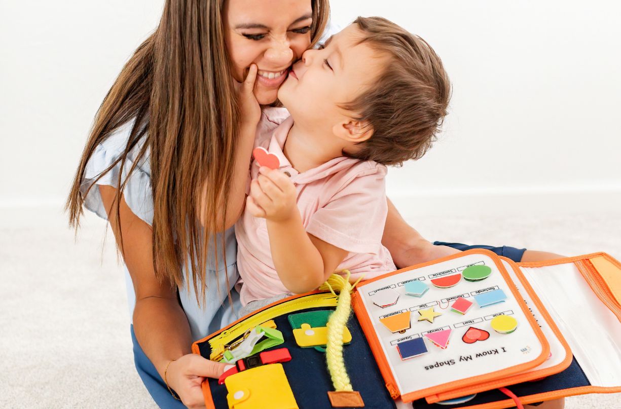 Boy kissing his mom while looking at the shapes sorting page of Milleli busy board - toddler travel toy for car and airplane